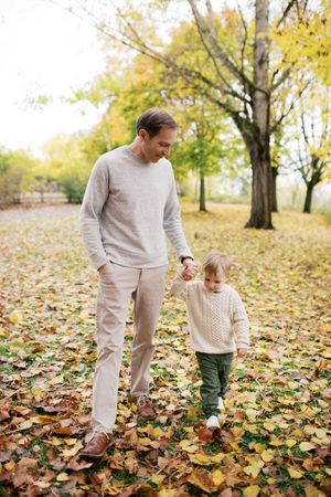 Parent and toddler play together in autumn leaves as golden trees create a warm fall backdrop in the park.
