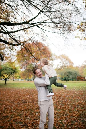 Playful autumn scene in a park with fallen leaves as father lifts child joyfully among orange foliage and bare trees.