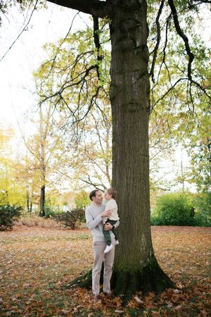 Person and dog embrace by a large tree trunk in autumn park with fallen leaves scattered on the ground.