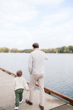 A parent and child in beige and green clothing stand together by a lake on an autumn afternoon.