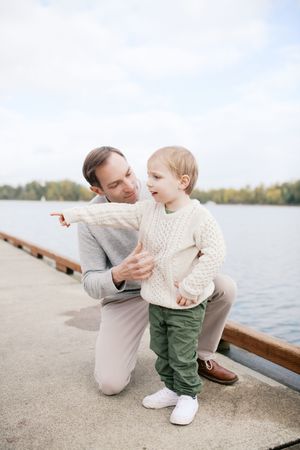 A parent and child share a tender moment on a dock by a lake, both wearing cozy sweaters on a cloudy day.