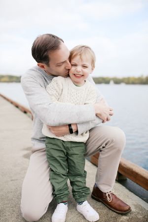 A parent and child share a tender embracing moment beside a lake while sitting on a wooden dock.