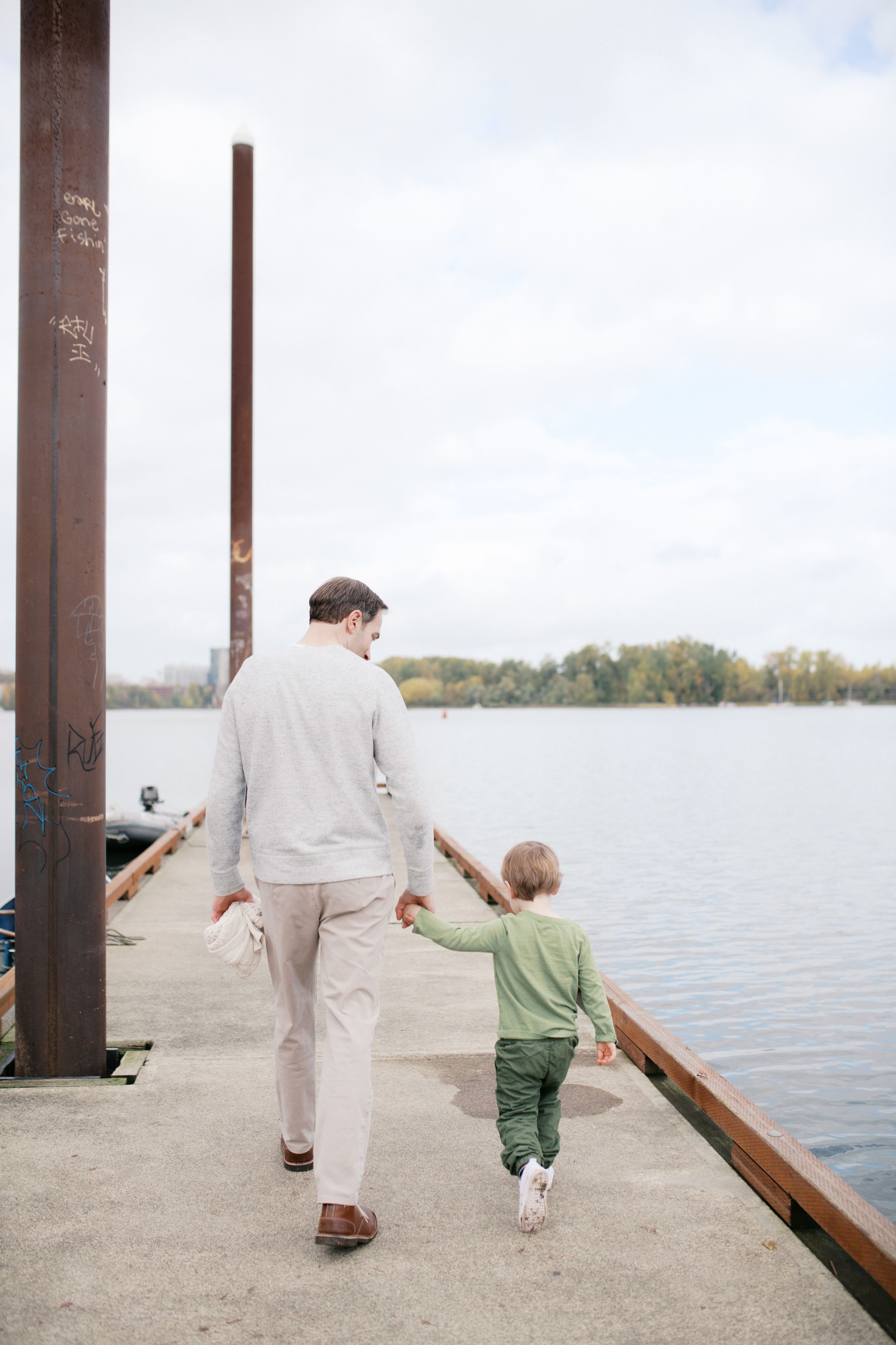 Adult and child holding hands while walking along a waterfront pier on a cloudy day.