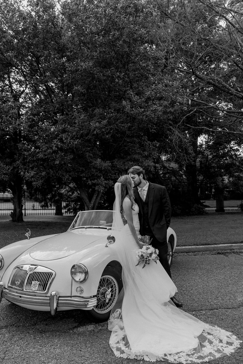 Classic white MG convertible parked under trees serves as backdrop for elegant wedding photograph.