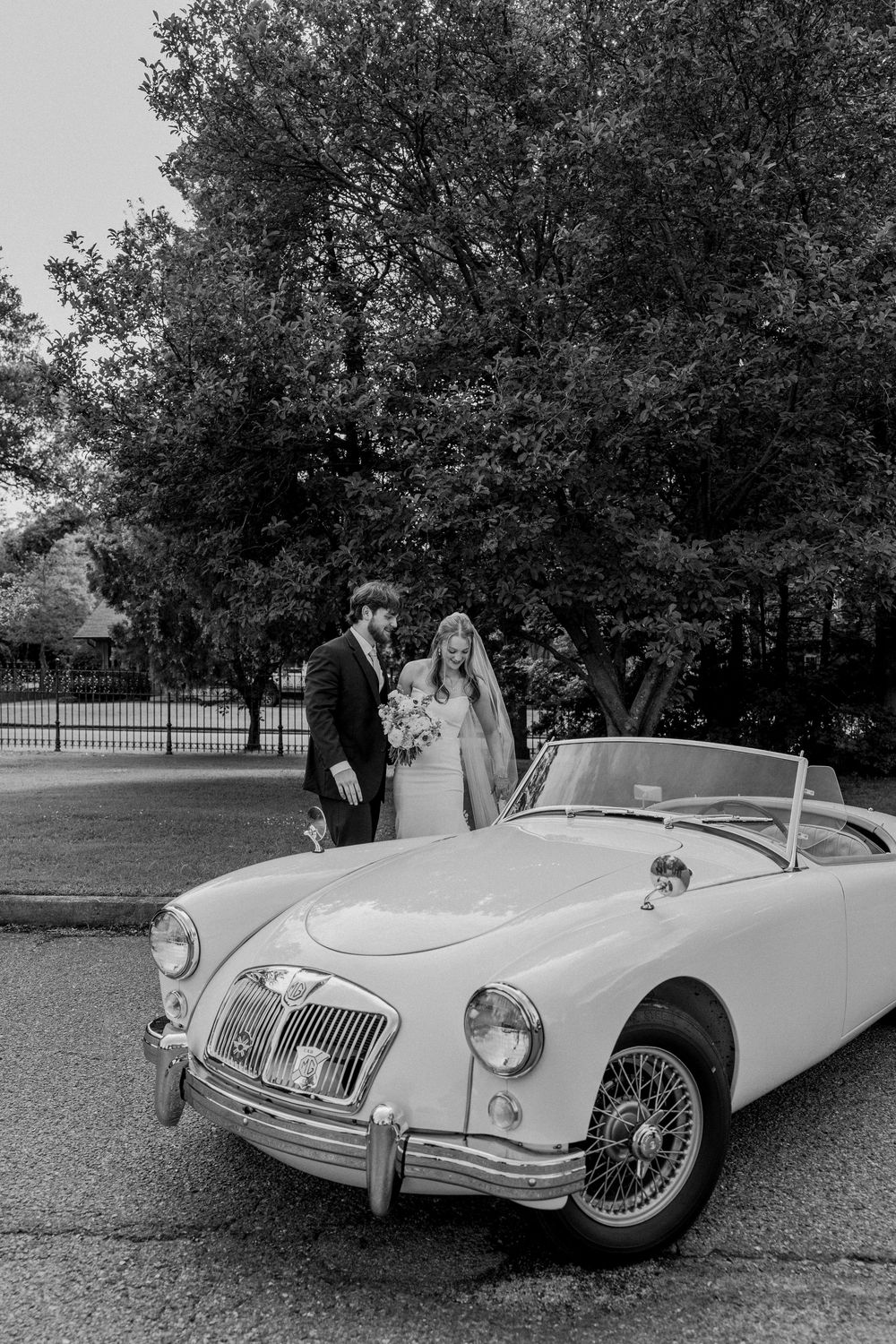 A classic vintage white MGA convertible car is parked in front of lush trees on a driveway during a special occasion.