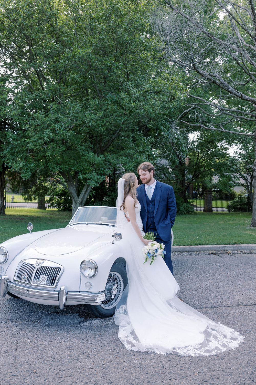 A vintage white MG sports car decorated for a wedding ceremony under green leafy trees.