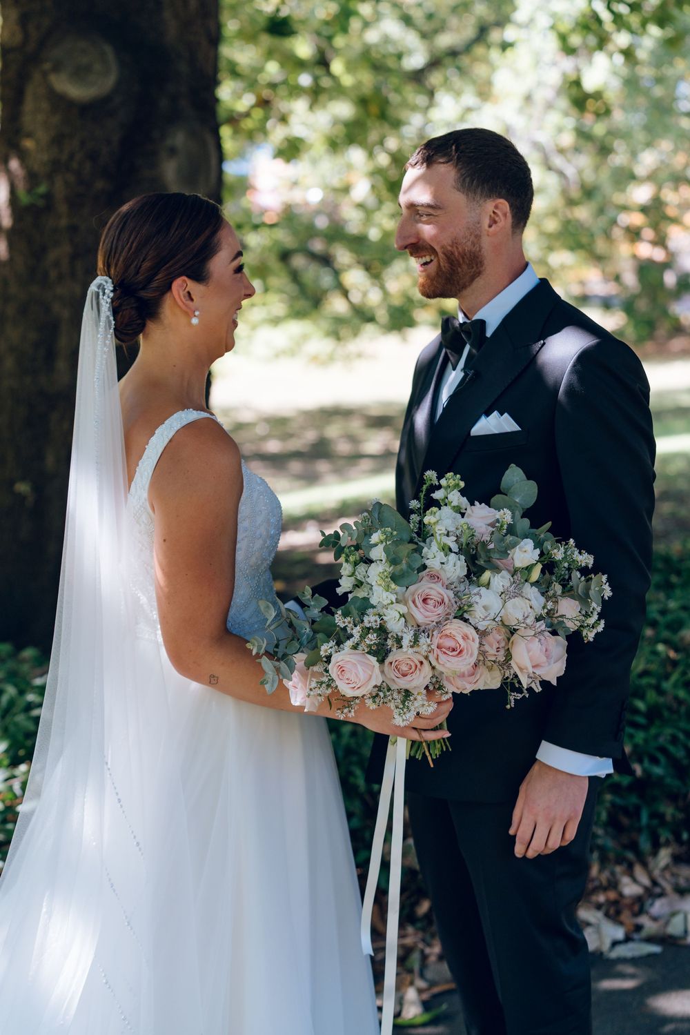 A bride and groom share a joyful moment outdoors while holding a beautiful bouquet of pink and white flowers.