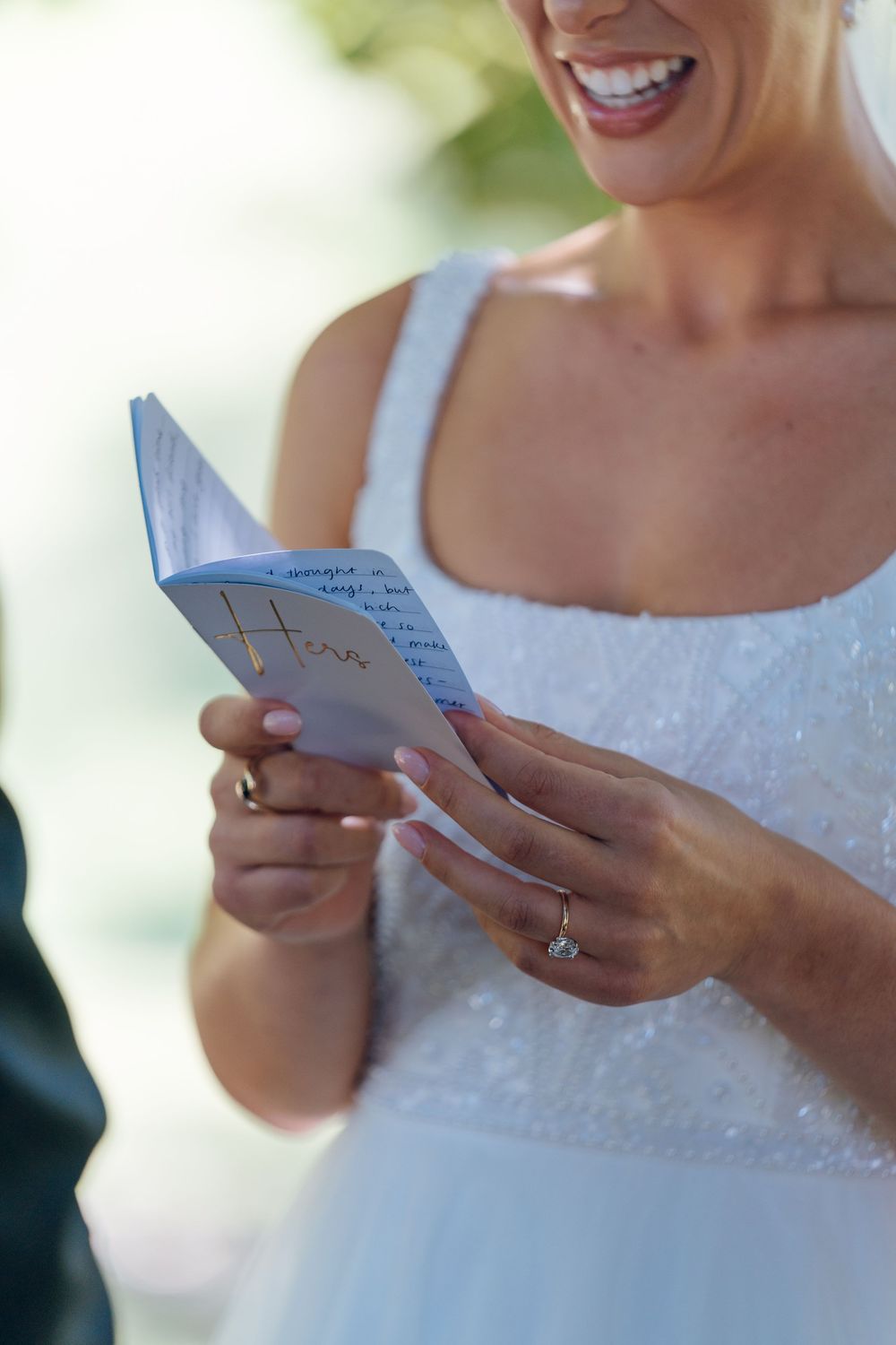 Close-up of a bride smiling while reading handwritten wedding vows during an outdoor ceremony.