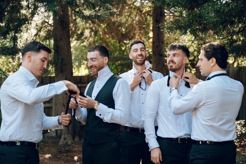 Groomsmen in white shirts and dark vests laughing and joking together outdoors.
