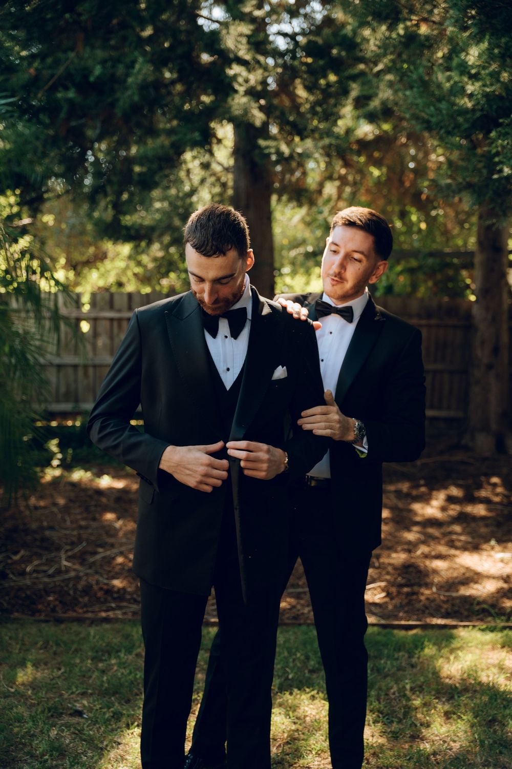 Two people in black tuxedos adjusting their bowties in a sun-dappled garden.