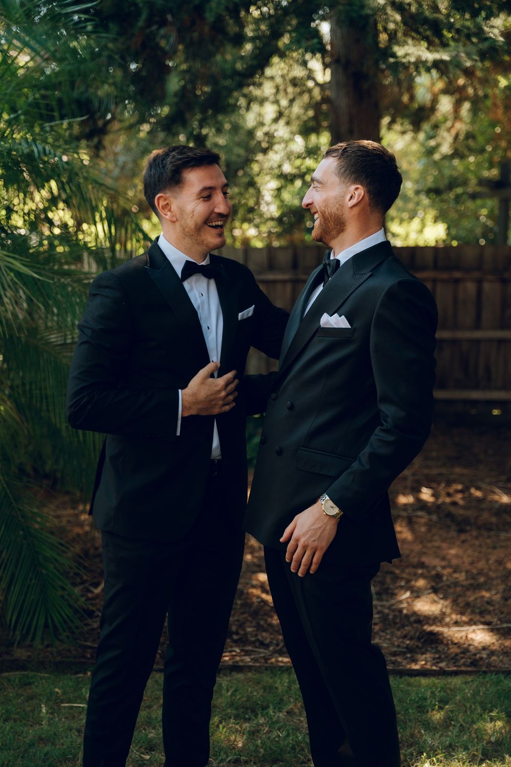 Two people in black tuxedos sharing a lighthearted moment outdoors.