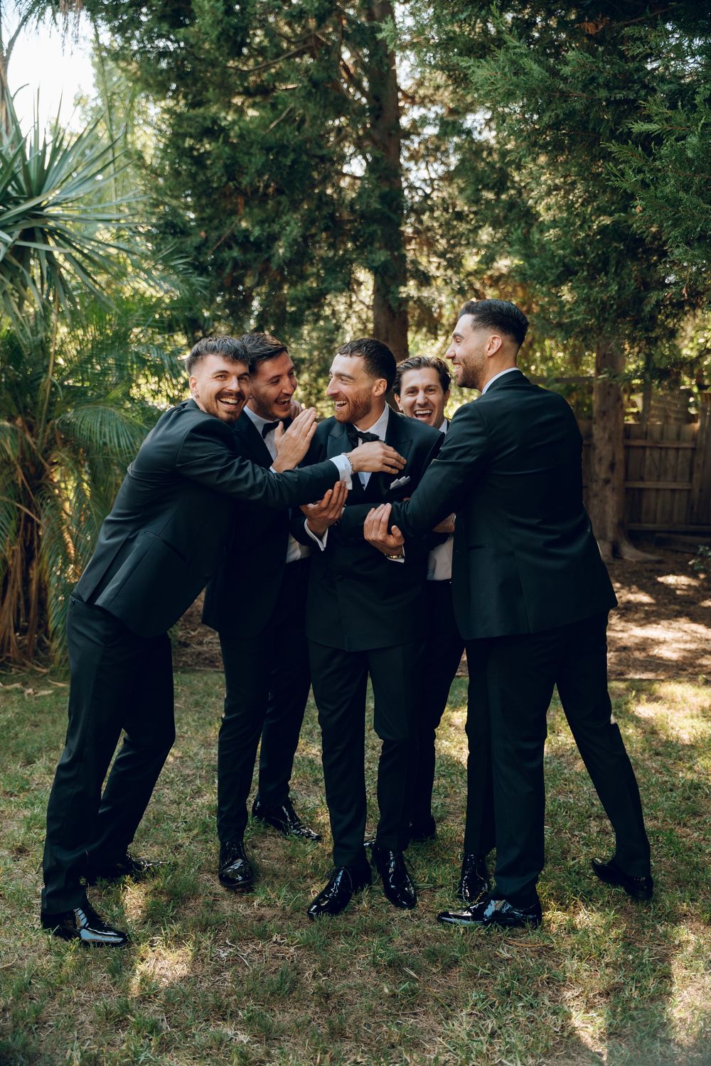 Group of formally dressed people hugging and laughing together in a garden setting.