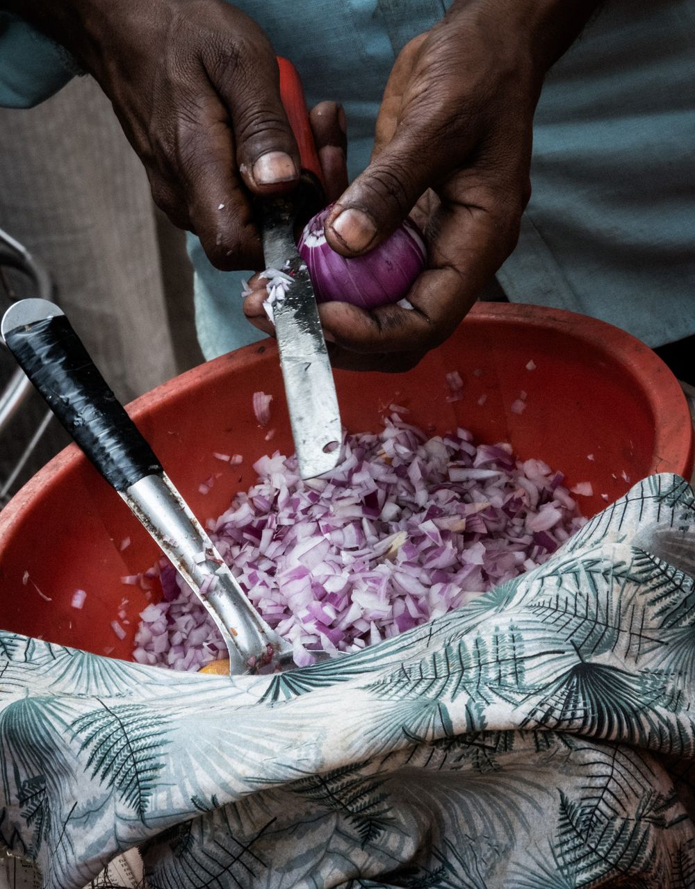 Hands chopping red onions with two knives into a large orange bowl.