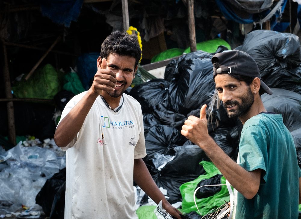 Two smiling men giving thumbs up near bags of collected recyclable waste.