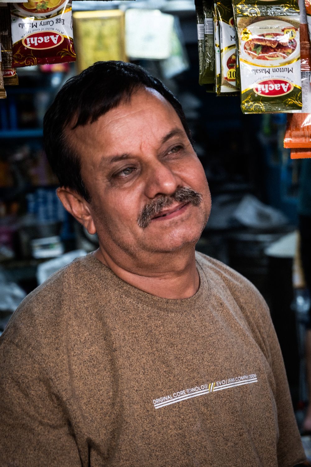 A mustachioed shopkeeper smiles in his store surrounded by spice packets.