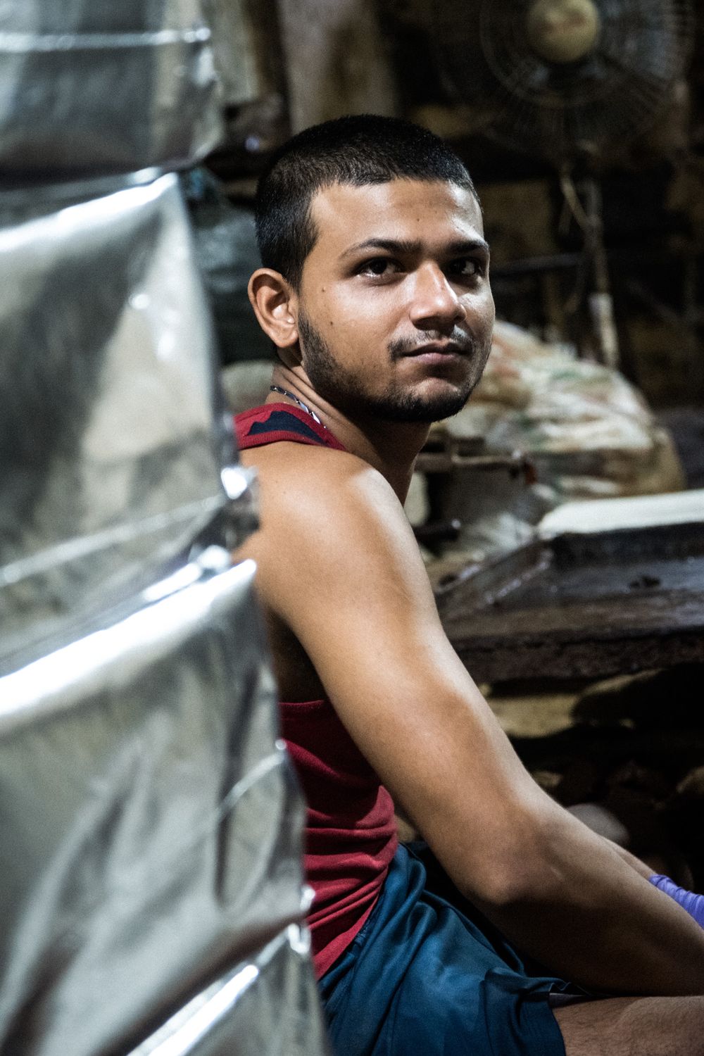 Young Indian man in red sleeveless shirt sitting in a workshop surrounded by metal materials.