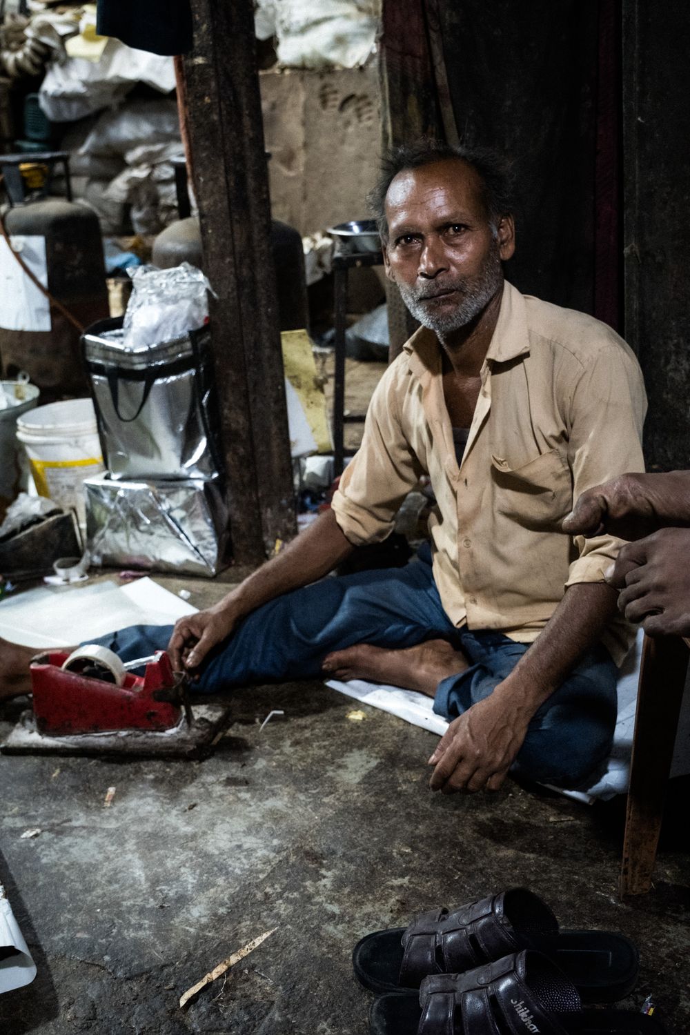 Elderly Indian man sitting cross-legged on floor in cluttered workshop holding a tool.