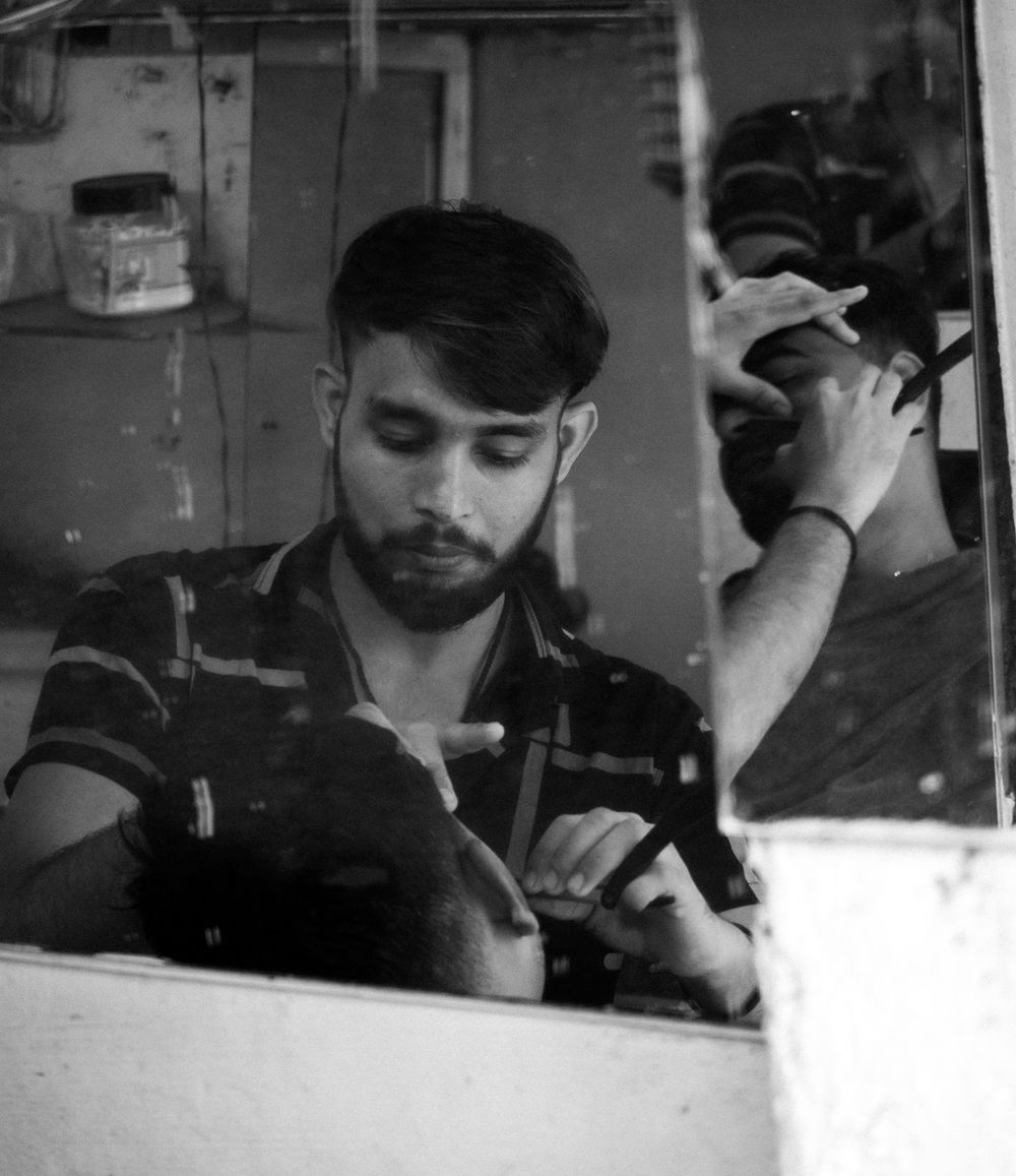 Black and white photo of bearded barber cutting hair in a small barbershop.