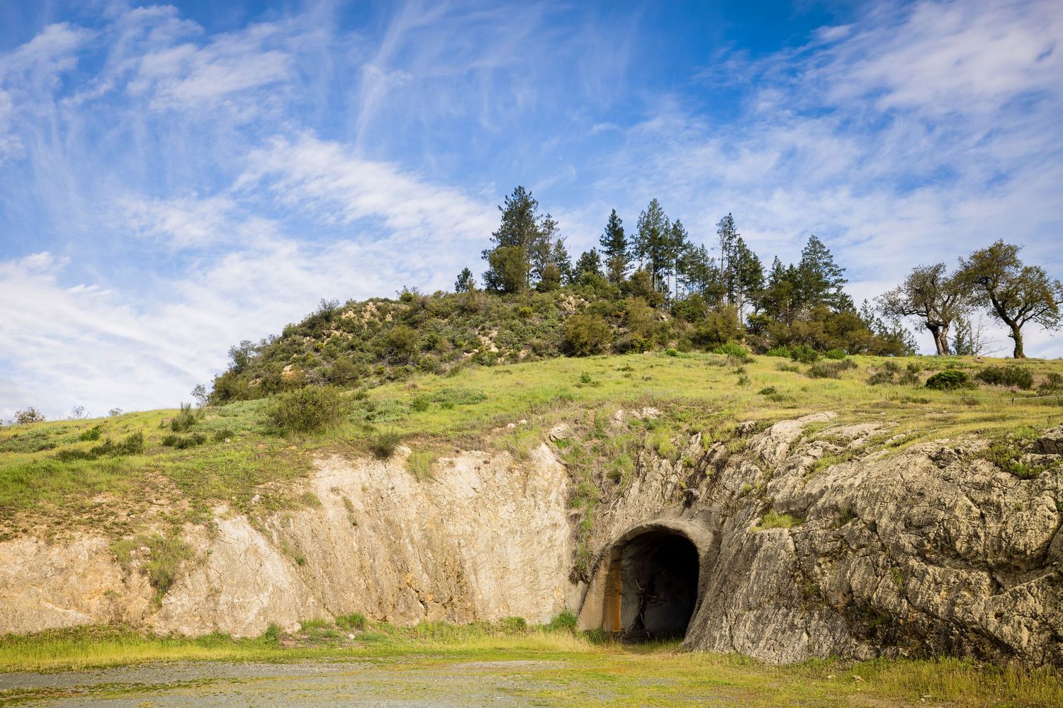 A tunnel entrance carved into a rocky hillside with evergreen trees on top against a bright blue cloudy sky.