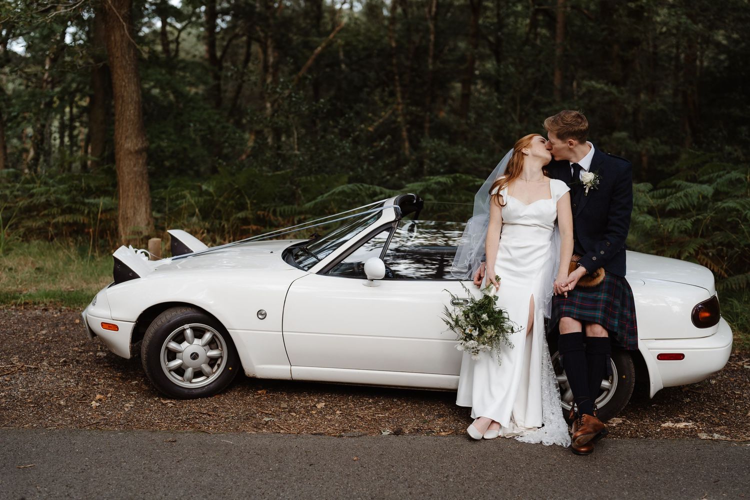A white convertible Mazda Miata parked in a wooded area during a wedding photoshoot.