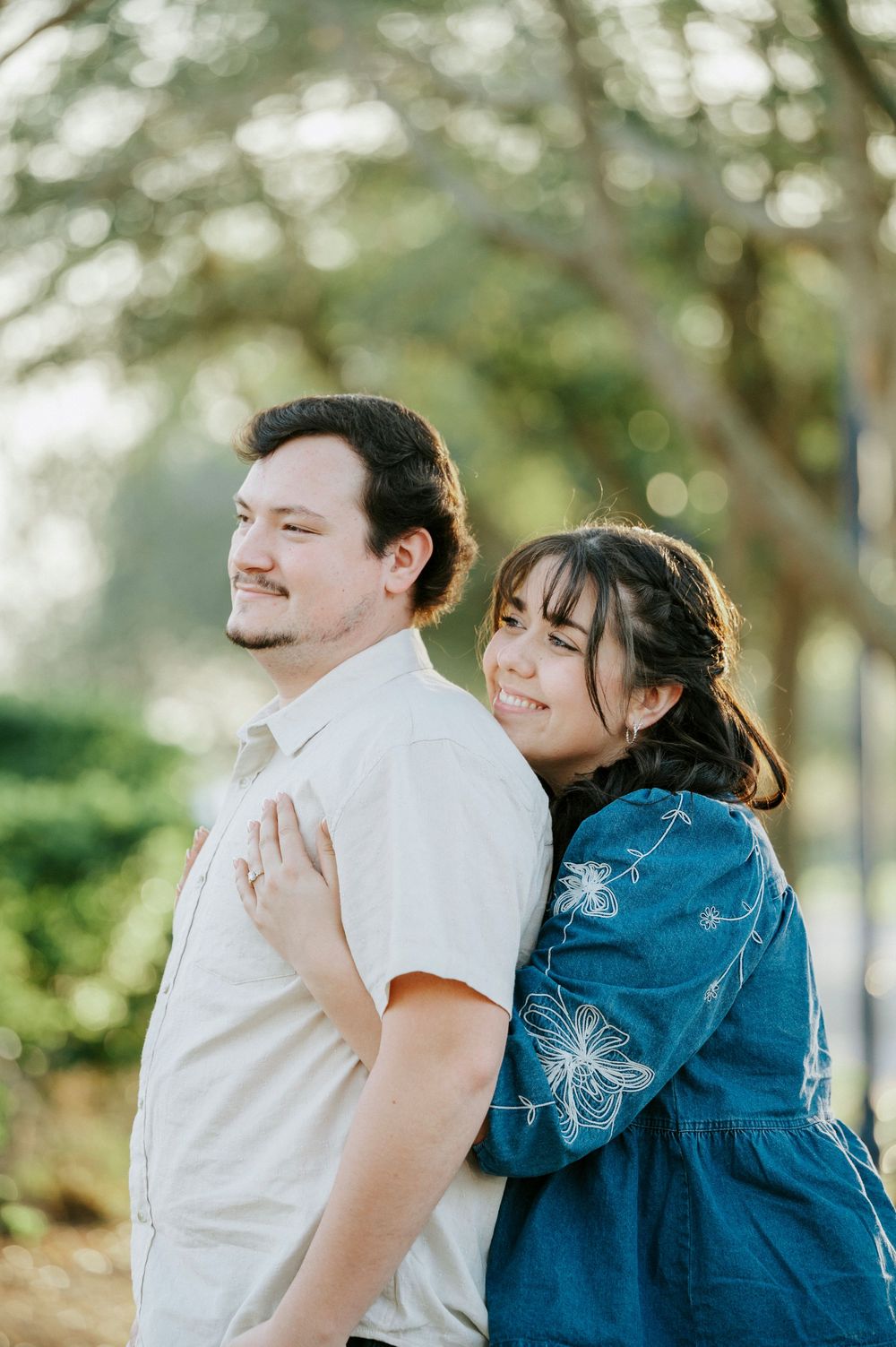 A couple shares a tender embrace in a sun-dappled outdoor setting, with one wearing a denim jacket.