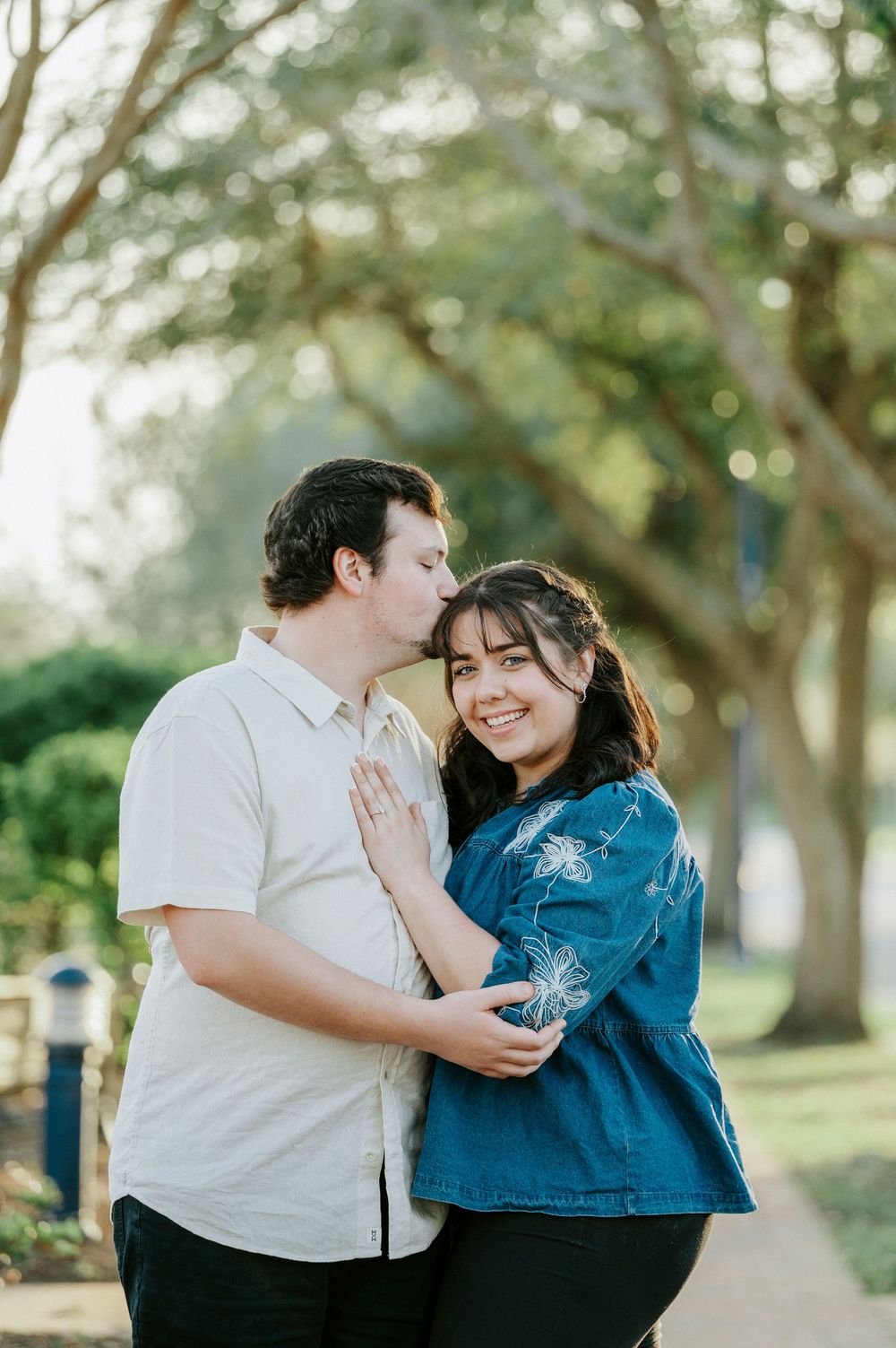 Couple in casual attire sharing tender moments outdoors in a park setting with trees in background.