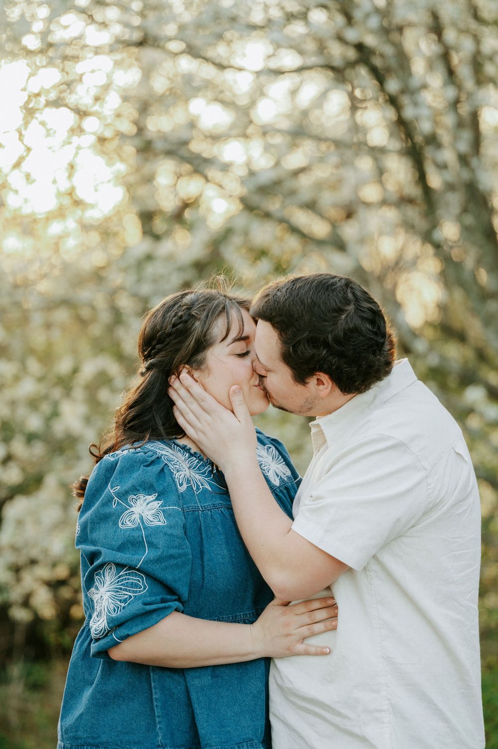 A romantic couple shares a tender kiss outdoors in front of blooming spring trees with soft bokeh lighting.
