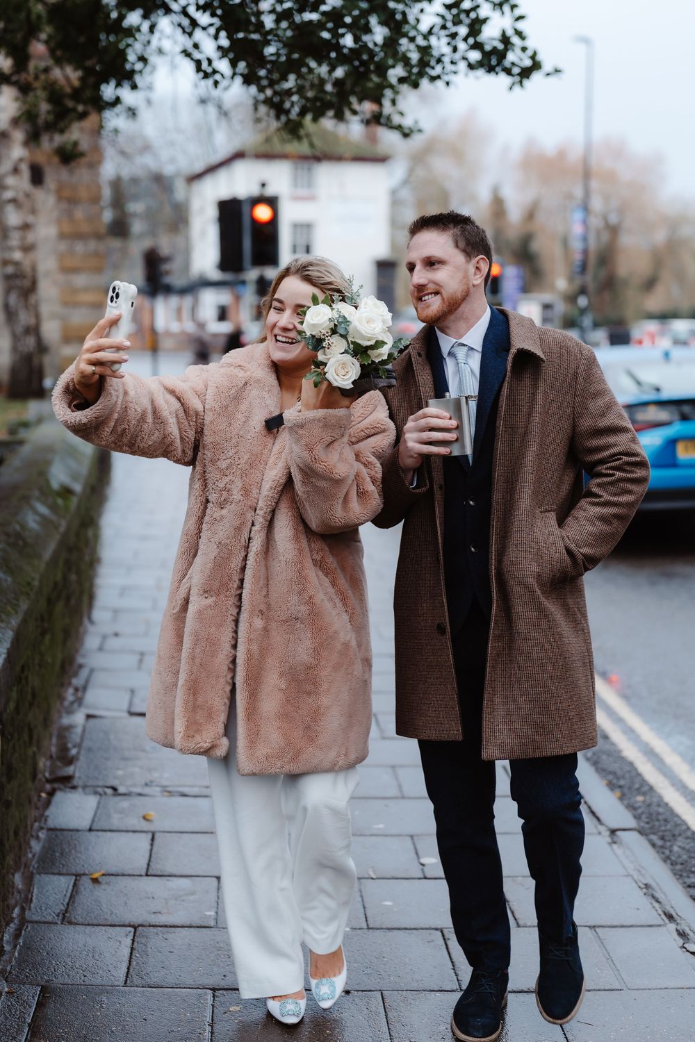 Couple in brown coats taking selfies together on urban sidewalk.