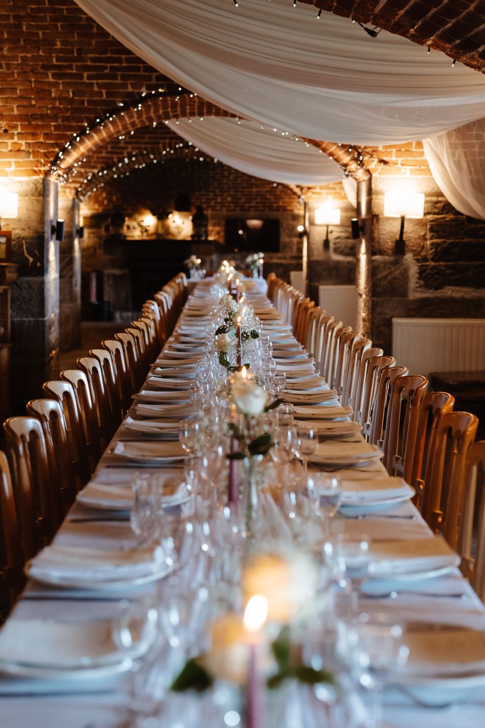 Elegant wedding reception table setting with white draping and string lights in a brick venue.