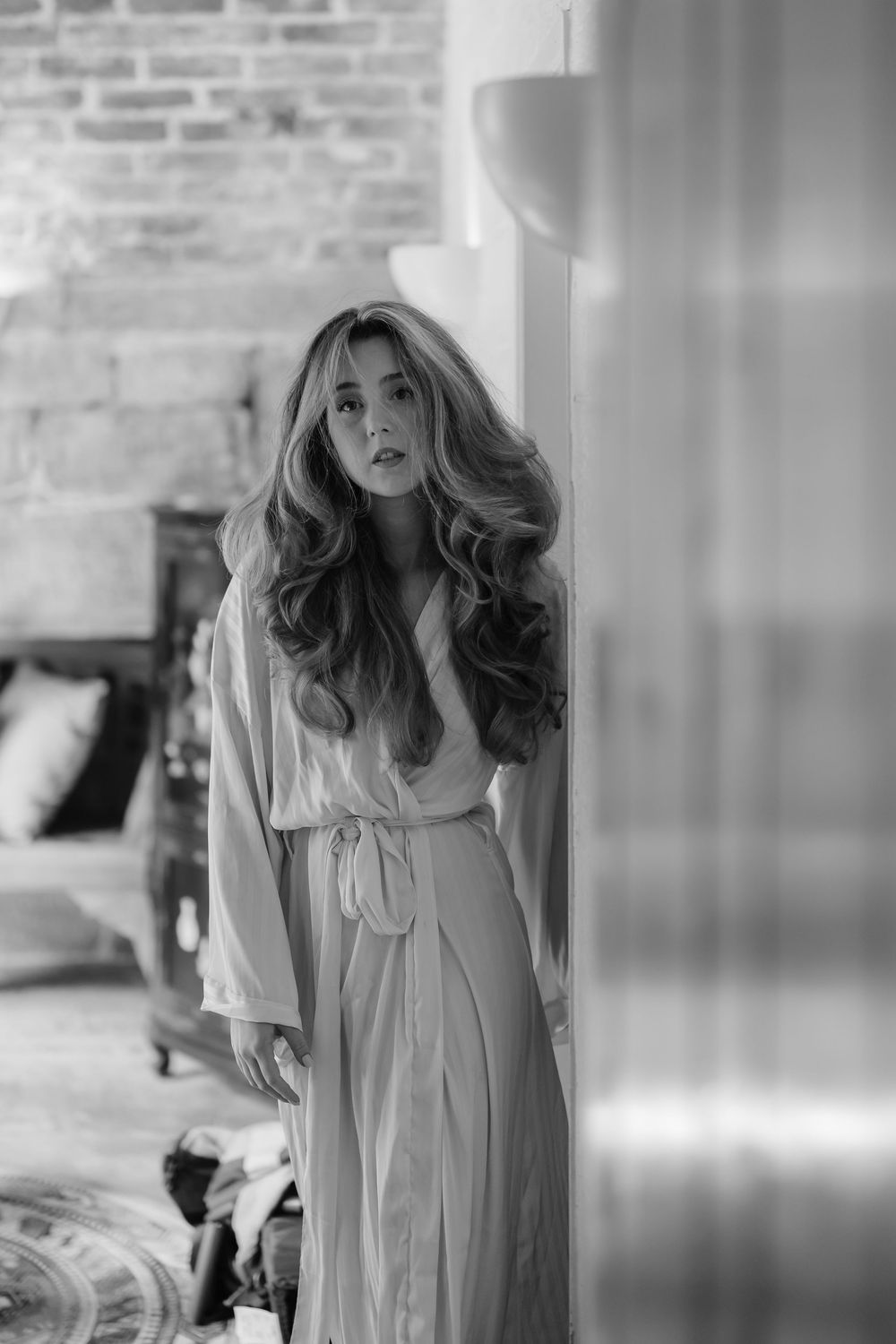 Black and white portrait of a woman in a flowing robe by a brick wall.