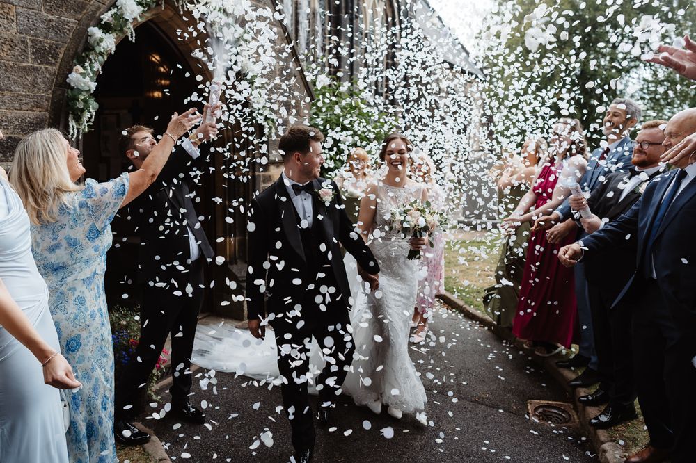 Wedding guests throw white confetti as newlyweds exit a stone church.