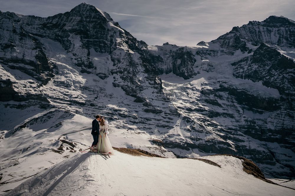 A couple poses on a snowy mountain peak with dramatic alpine peaks in the background.