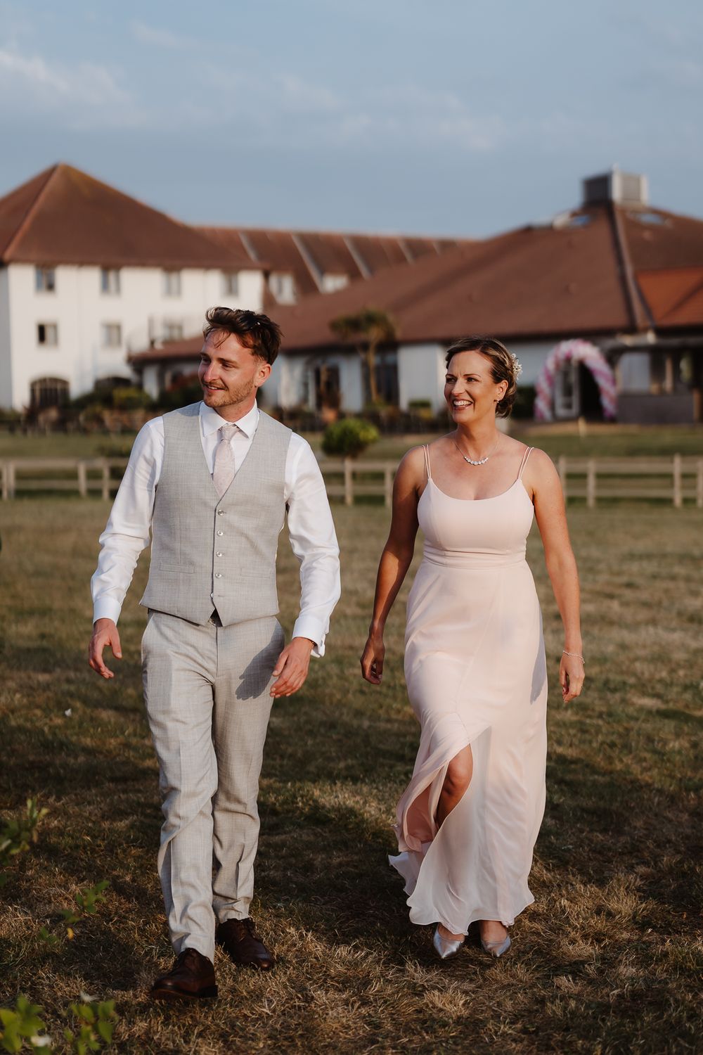 Couple walking together in formal attire across a grassy field with houses in background.