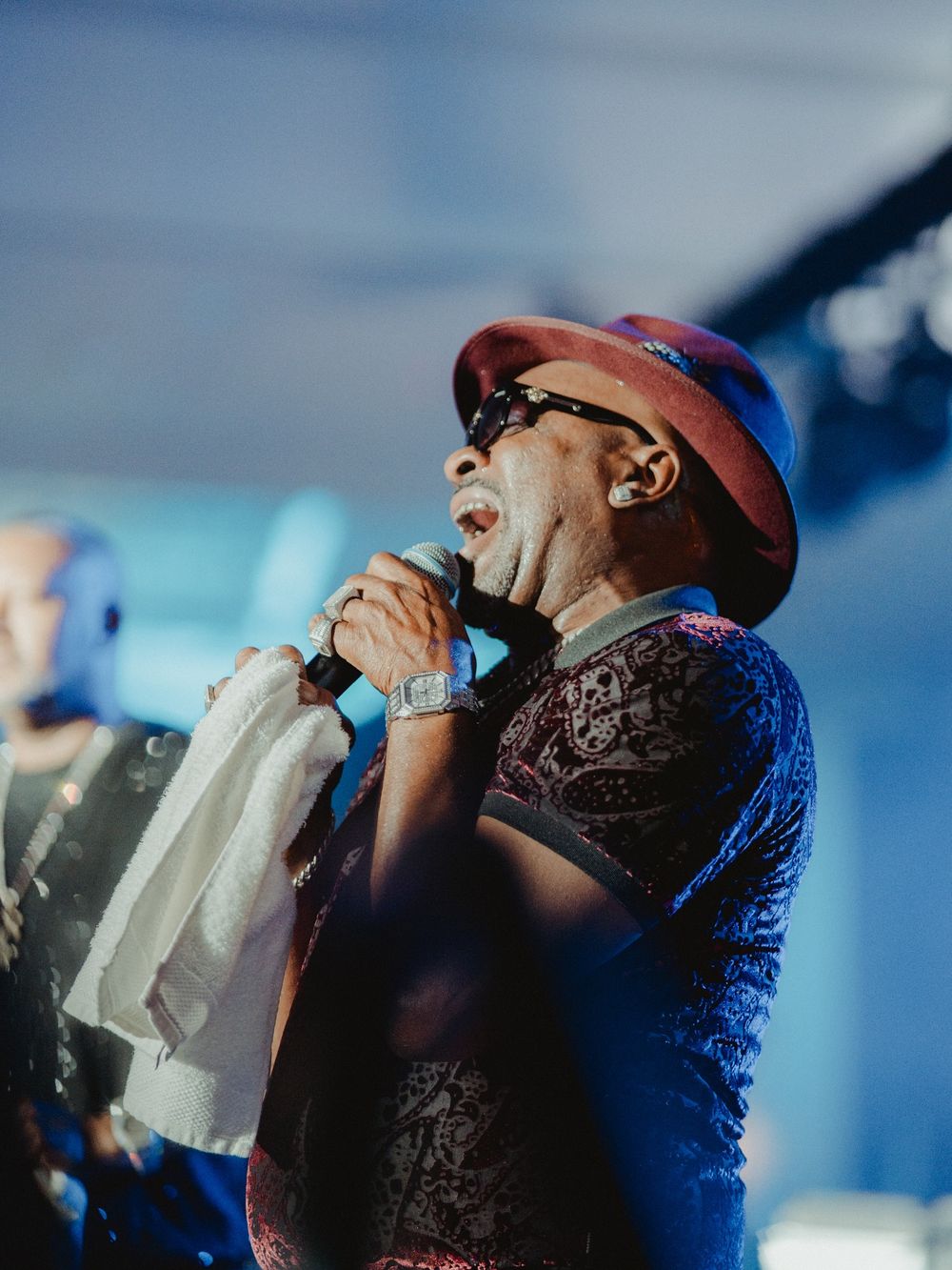 A performer in a black shirt and brown hat sings passionately into a microphone against a blue-lit stage background.