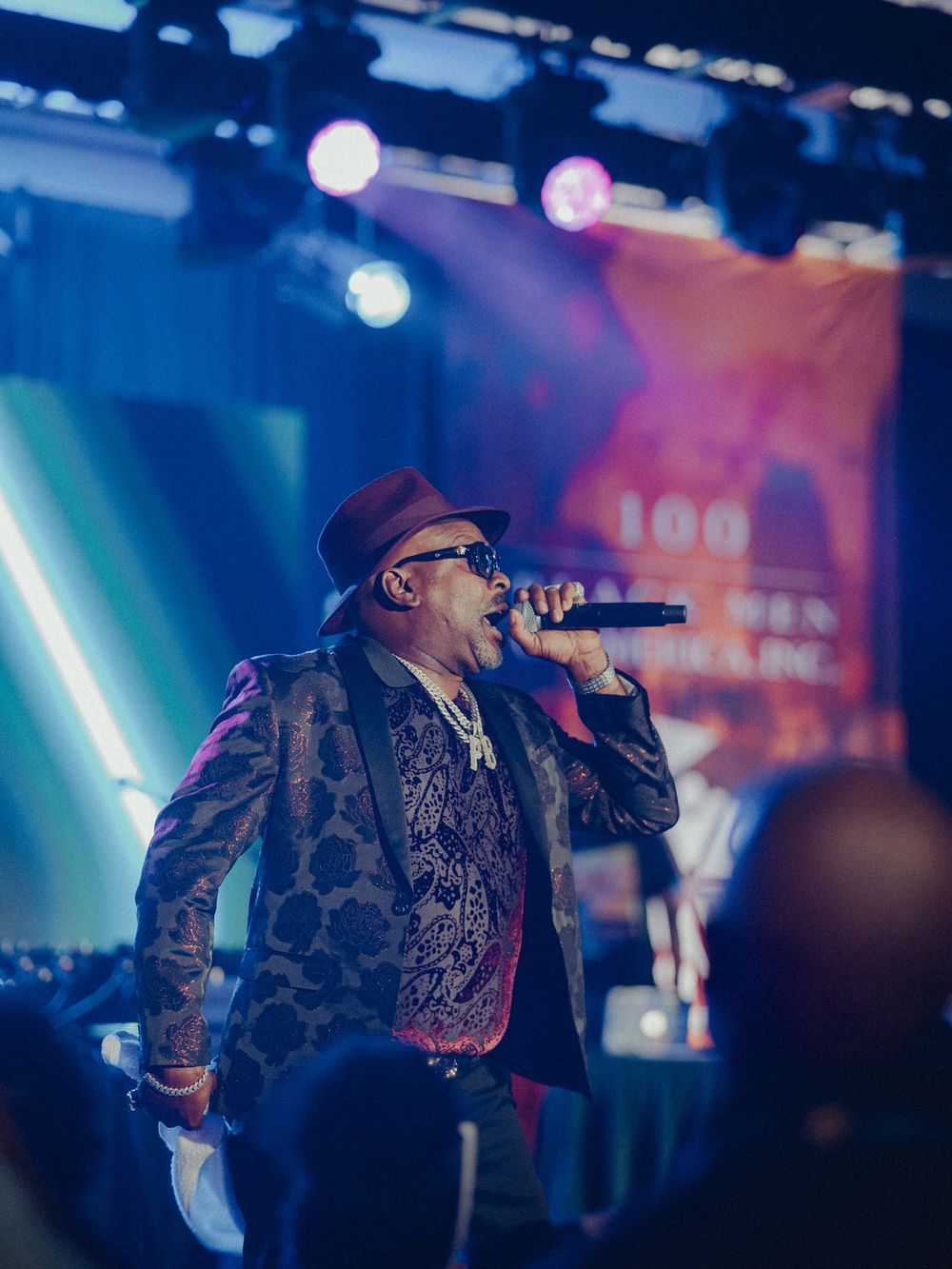 Performer in sunglasses and patterned jacket energetically singing on stage with colorful blue and pink lighting effects.