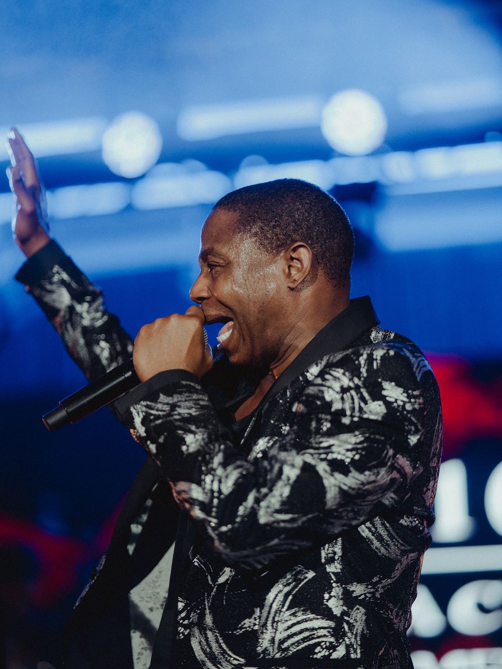 Performer in black and gray patterned jacket sings passionately on stage with blue lighting effects in background.