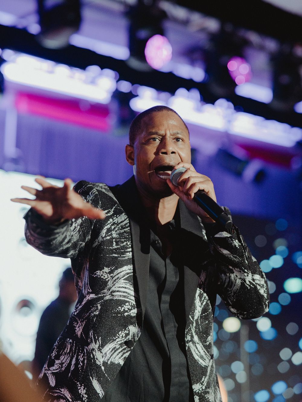 A performer in a black and silver patterned jacket sings into a microphone on stage with blue lights in the background.