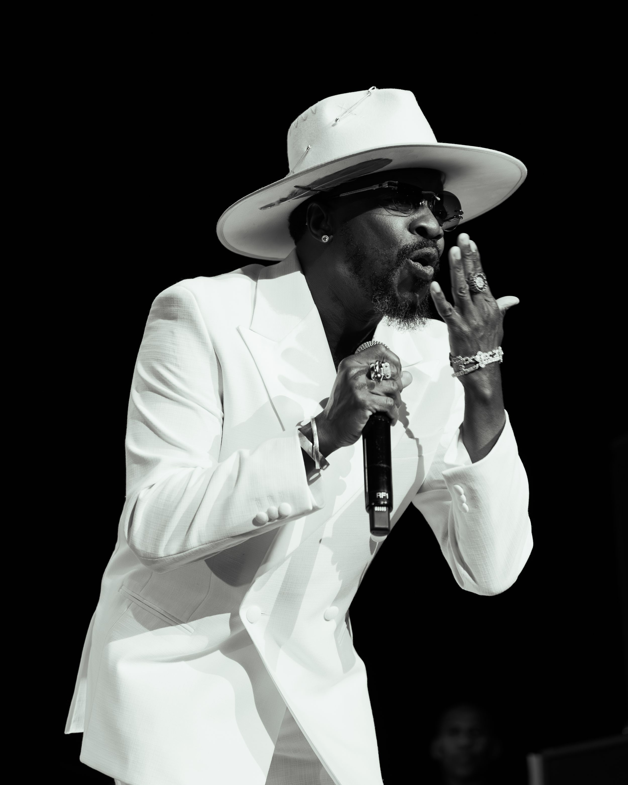 A performer in a white suit and cowboy hat sings passionately into a microphone on a dark stage.