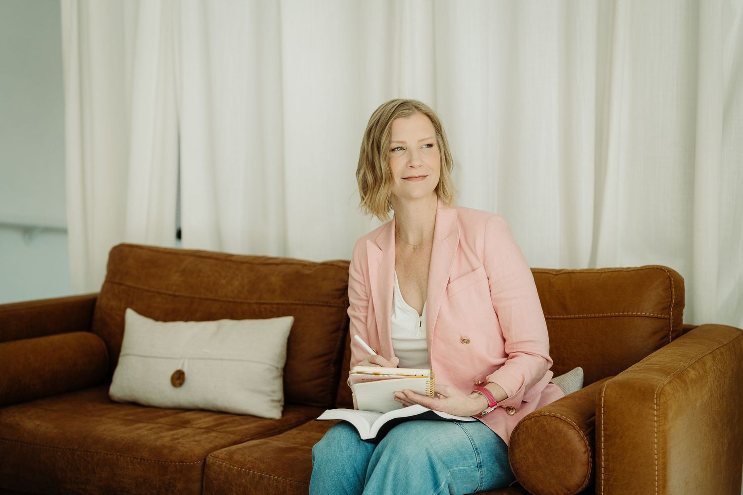 Person in pink blazer and blue pants sits on brown leather couch with book and decorative pillow in background.