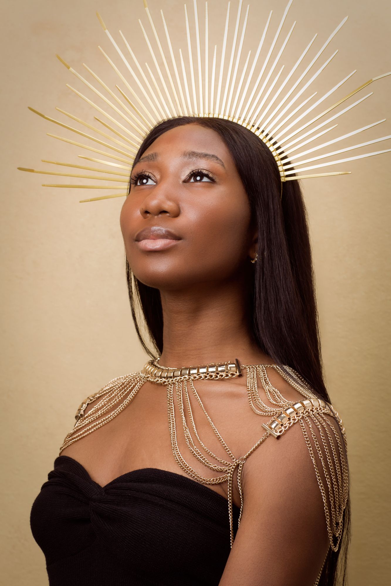 Studio portrait with gold sunburst crown and draped chain shoulder jewelry against beige background.