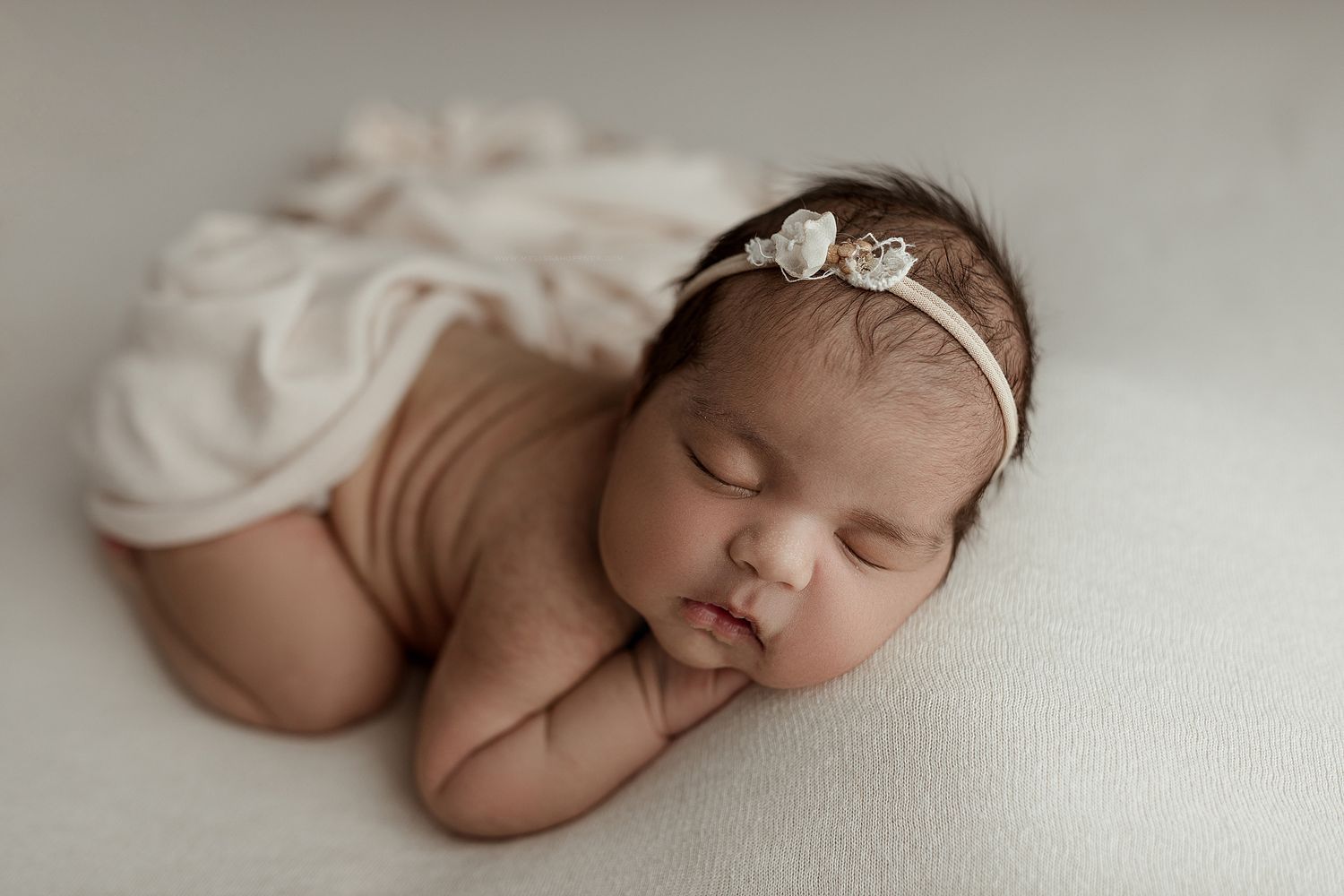 A sleeping newborn baby wearing a white headband lies peacefully on a cream-colored blanket in a professional photo setting.