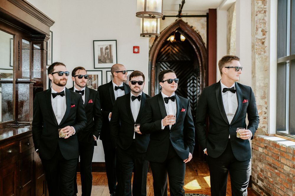 Groomsmen in black tuxedos and bowties share a toast with drinks in an elegant venue hallway.