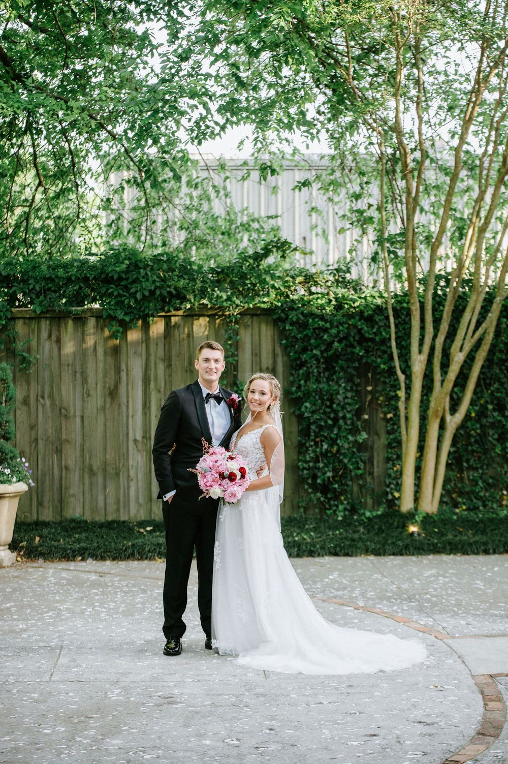 A wedding couple poses together in a garden setting with a wooden fence and green trees in the background.