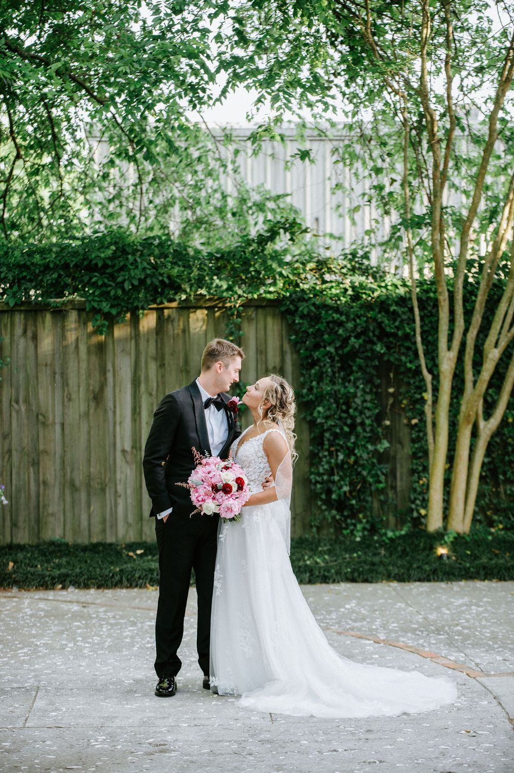 A couple in formal wedding attire share an intimate moment in a garden setting with a pink bouquet.