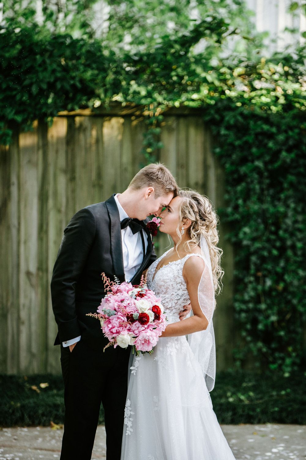 A romantic wedding couple shares intimate moments in a garden setting with pink floral bouquet details.