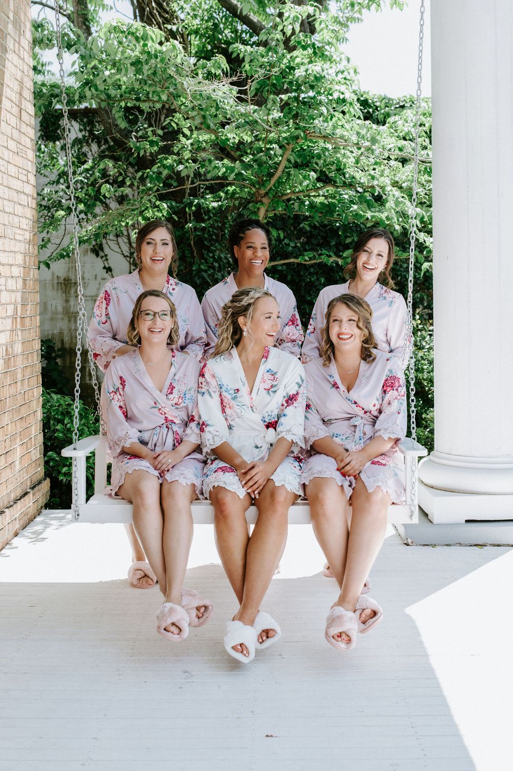 A group in matching floral robes sits together on white porch steps during a wedding day preparation.