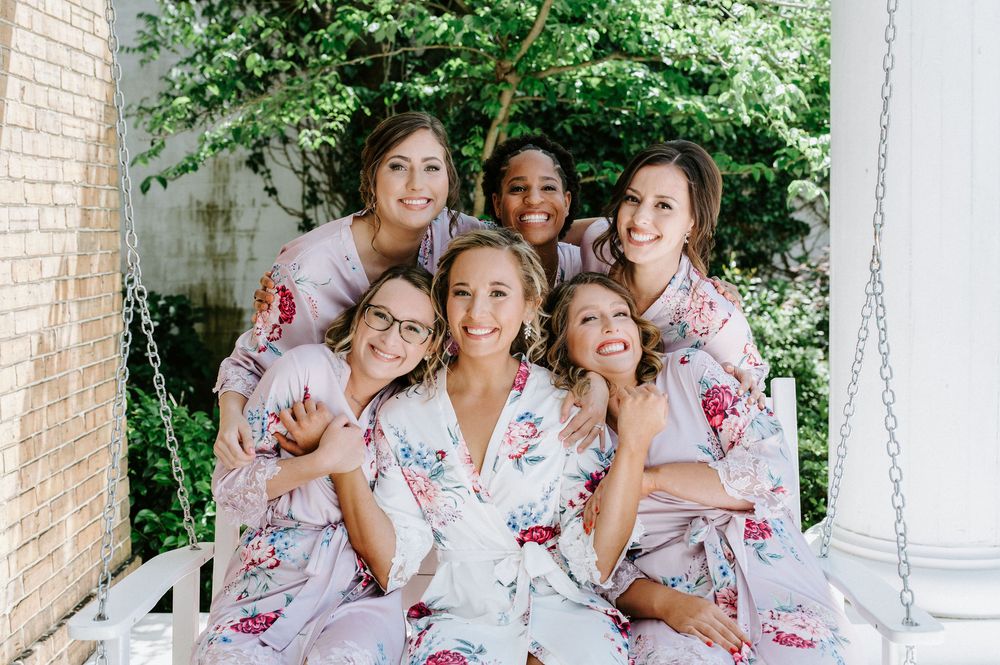 Bridesmaids in floral robes pose together on a white porch during wedding preparations.