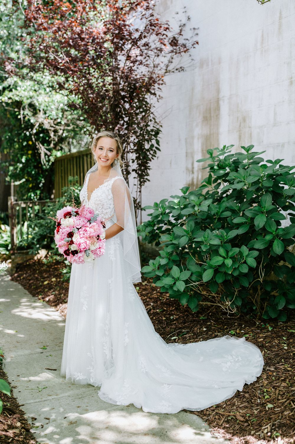 A person in a flowing white lace wedding gown holds a vibrant pink bouquet while standing in a garden setting.