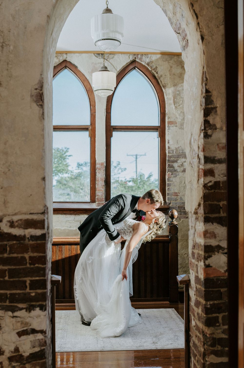 Romantic wedding couple dips and kisses in front of arched windows in rustic brick venue.