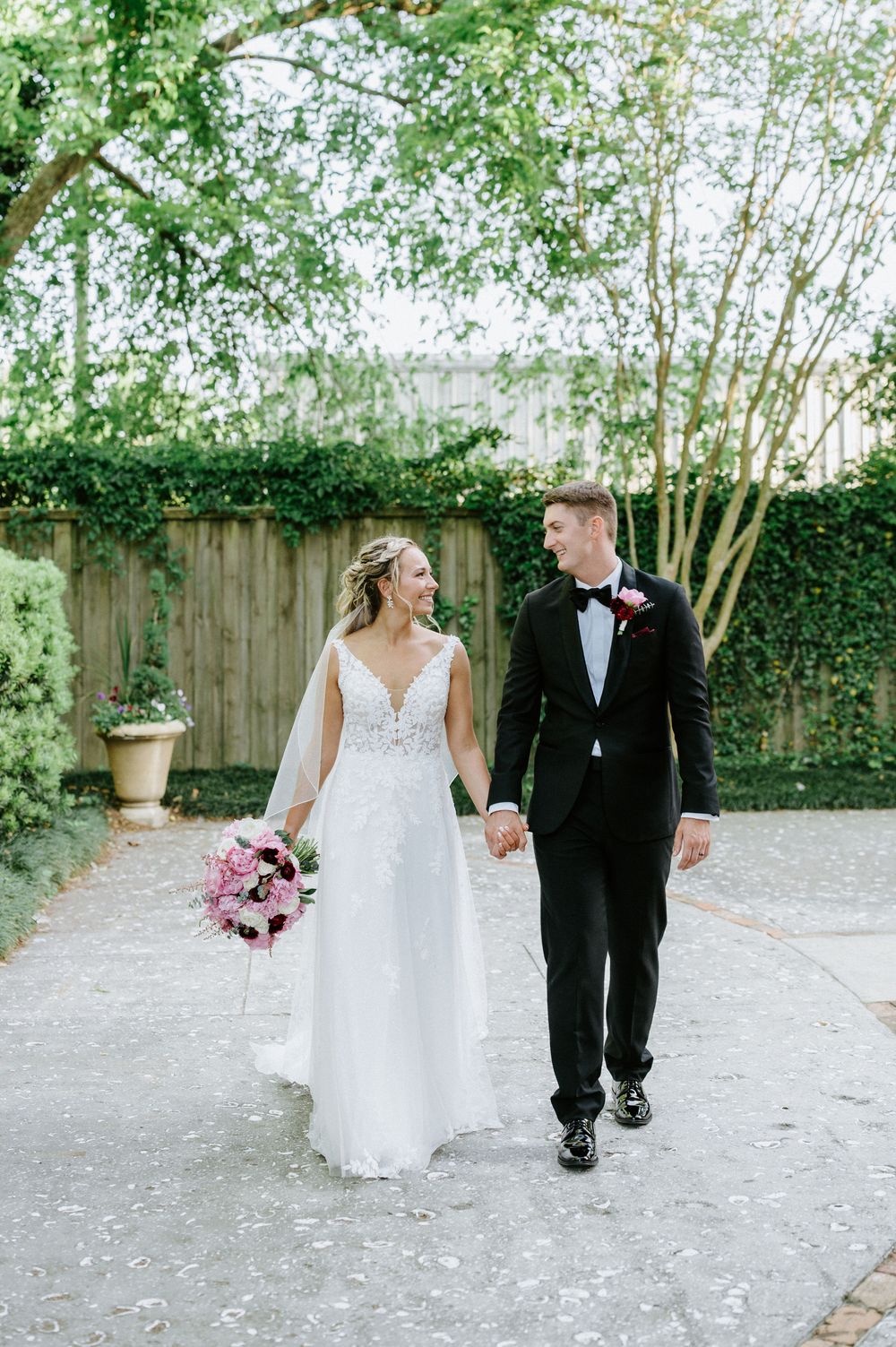 A couple walks hand in hand on their wedding day in a garden setting with greenery and a wooden fence.